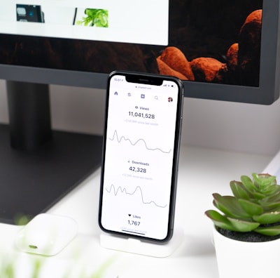 A smartphone displays analytics data with graphs showing views, downloads, and likes. It is resting vertically on a white dock next to a small potted succulent plant. In the background, a large computer monitor is partially visible, along with a closed laptop and a white wireless earbud case.