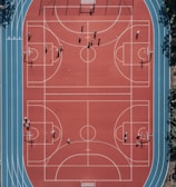 An aerial view of a large, vibrant sports complex featuring a red basketball court with multiple hoops and a surrounding blue running track. Several people are scattered across the area, engaged in various activities.