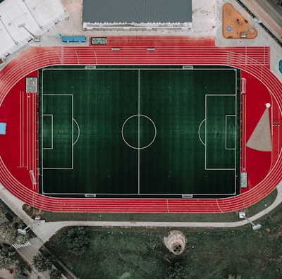 An aerial view of a large sports complex featuring a central green football field surrounded by a red running track. Adjacent to the track, there is a parking area, buildings, and pathways. Nearby, there are additional sports courts in different colors, including a blue swimming pool and orange basketball courts.