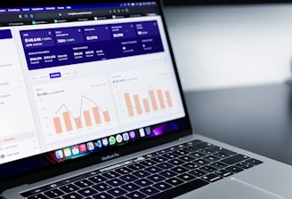 A laptop with an open screen displaying dashboard analytics featuring charts, graphs, and numerical data related to sales and subscriptions. The interface is predominantly dark-themed with sections highlighting key performance metrics. The device shown is a MacBook Pro, indicated by the keyboard layout and branding.