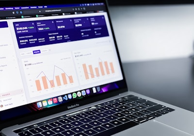 A laptop with an open screen displaying dashboard analytics featuring charts, graphs, and numerical data related to sales and subscriptions. The interface is predominantly dark-themed with sections highlighting key performance metrics. The device shown is a MacBook Pro, indicated by the keyboard layout and branding.